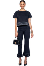 A woman modeling a black two-piece outfit consisting of a short-sleeve top with a textured horizontal band at the hem and matching black cropped pants with ruffled cuffs, holding a small black handbag against a light gray studio background.