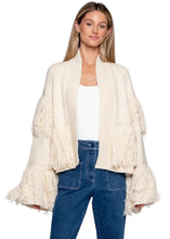 A woman modeling a cream-colored open-front knit cardigan with long fringe on the sleeves and hem, worn over a white top and blue jeans against a light gray studio background.
