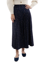 A model wearing a navy blue pleated midi skirt with a small beige floral pattern, styled with a cream cardigan and navy blue pointed flats.