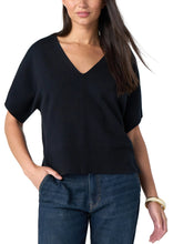 A woman modeling a black short-sleeve V-neck knit sweater with relaxed fit, paired with dark blue jeans, against a light gray studio background.